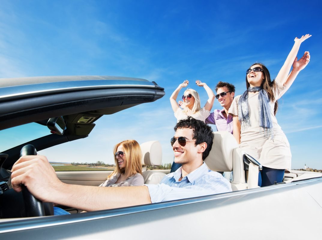 Side view of a group of young cheerful people with raised hands are enjoying in the car ride.