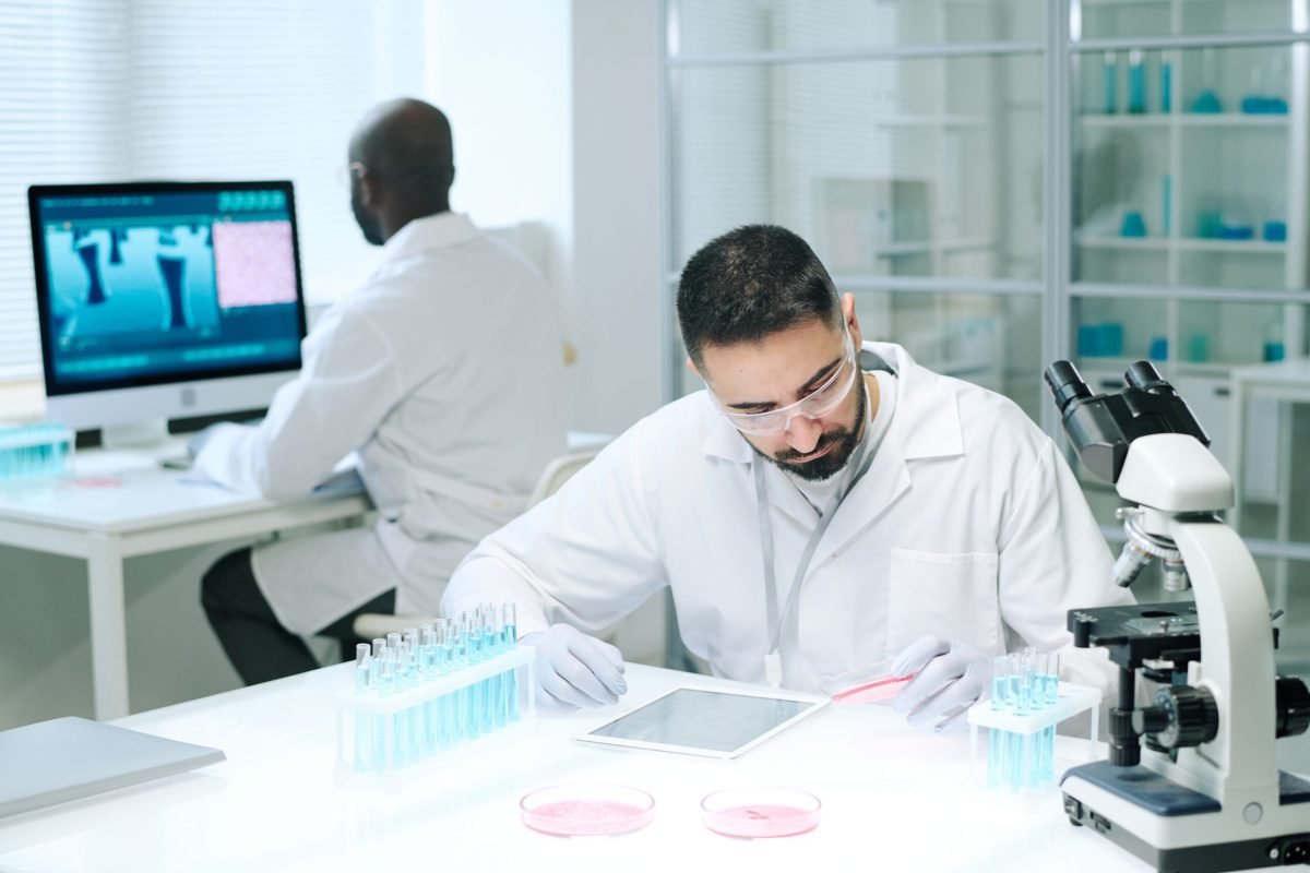 Young serious male chemist in whitecoat, protective gloves and eyeglasses studying substance in petri dish by workplace